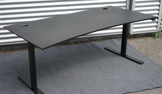 Dencon hæve-sænkebord 160x90 i sort linoleum med affasede kanter og elegant bue, set fra skrå vinkel med kabelbakker.
