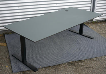 Dencon mørkegrå hæve-sænkebord set i skrå vinkel. Bordplade i mørkegrå linoleum med kabelgennemføring og sort kabelbakke, 200x90 cm.