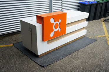 Receptionsbord set skråt fra højre side med hvid top, trædetaljer og orange skilt.