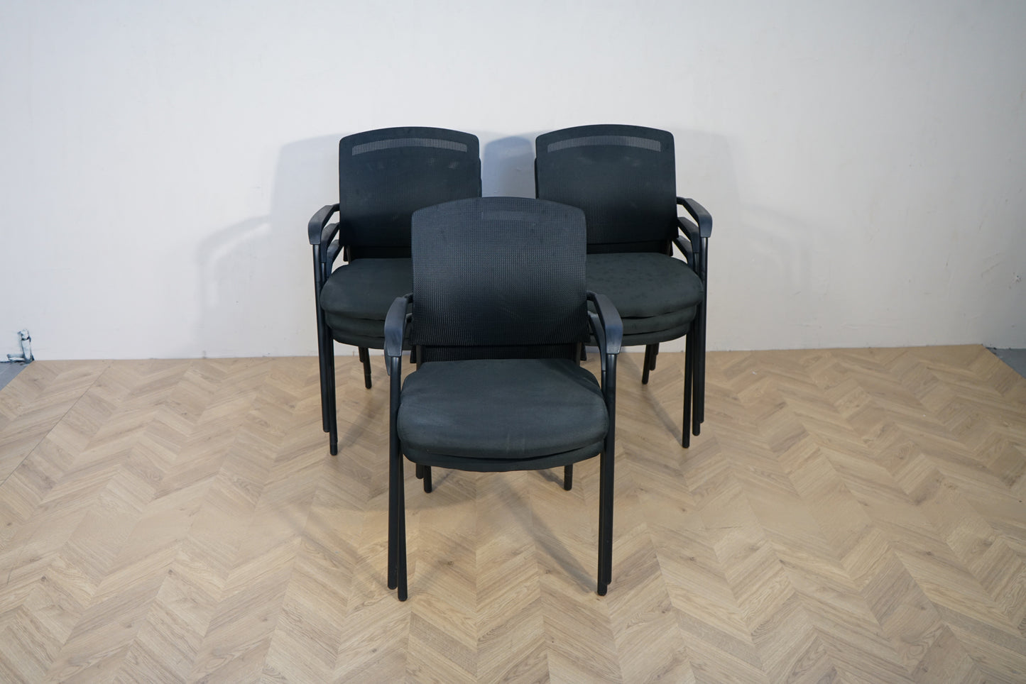 Sort stabelbare konferencestole med armlæn, mesh-ryg og polstrede sæder samlet set forfra.
